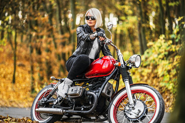 Obraz na płótnie Canvas Pretty blonde biker girl in sunglasses with red motorcycle on the road in the forest