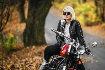 Naklejka na ściany i meble Pretty blonde biker girl in sunglasses with red motorcycle on the road in the forest