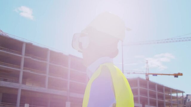 Professional builder in VR helmet standing in front of construction site and using virtual and augmented reality technologies. Office building and crane background. Real estate and investment.