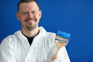 Portrait of paintbrush covered with blue paint. Smiling male worker in uniform on wall background. Professional interior designer. Renovation and construction site concept