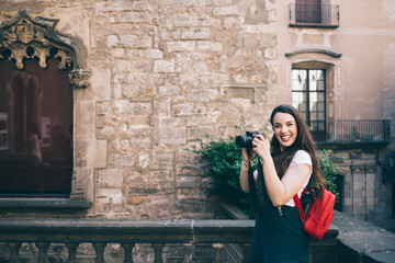 Cheerful caucasin female amateeur of photography enjoying free time on making pictures of urban setting on sunny day, smiling hipster girl tourist taking photos during trip excited with equipment