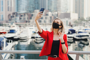 Young woman in red business suit wearing protective face mask of black color taking a selfie photo near yachts and skyscrapers in city center. Travel during Covid 19 or coronavirus pandemic