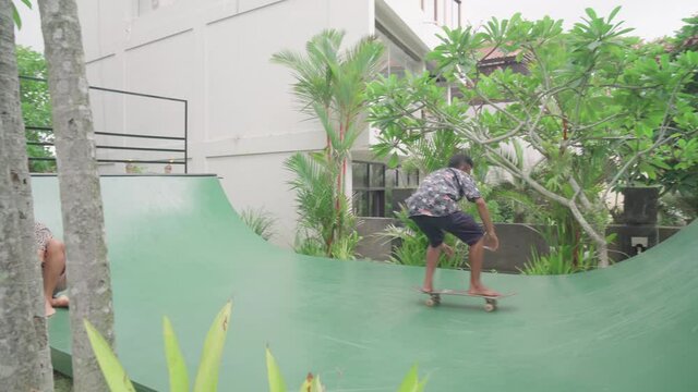 Young Asian man trying to ride skateboard while his friend playing with his little cute dog on halfpipe in backyard of villa, handheld wide shot