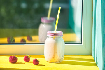 strawberry smoothie in a jar with drinking straws on a windowsill on a bright sunny day. ripe strawberries on a yellow background