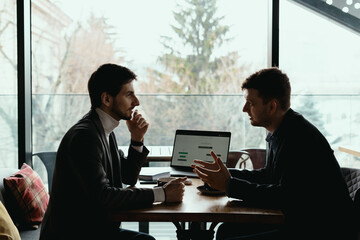 Two businessmen talking about new opportunities sitting with laptop at desk, planning project, considering business offer, sharing ideas