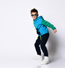 Little kid in sunglasses, colorful sport suit and sneakers. He performing a kick by his leg, posing isolated on white