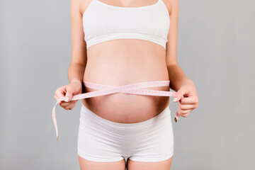 Close up of pregnant woman in white underwear measuring her growing tummy with a tape measure at gray background. Check a baby development. Copy space