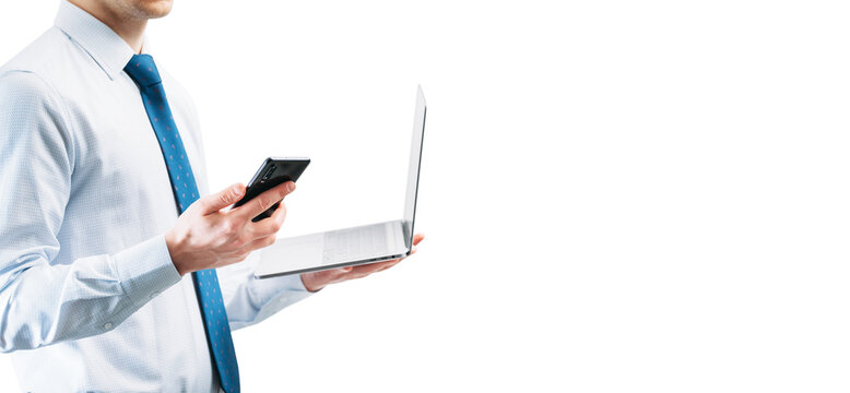 A guy in a light shirt with a tie on a white background looks at the phone screen while holding an open laptop. Panoramic photo with a place for an inscription..