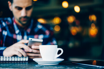 Select focus on white cup of tasty cappuccino with mock up area for advertising with brulled background of young hipster guy chatting online on smartphone device connected to high speed internet