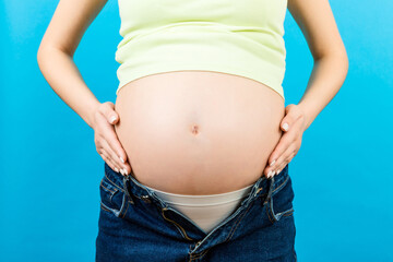 Close up of pregnant woman in opened jeans caressing her baby bump at colorful background with copy space. Waiting for a baby concept