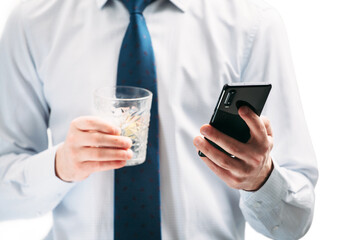 A guy in a white shirt and blue tie on a white background. A man looks at the phone screen and holds a glass with a cocktail.