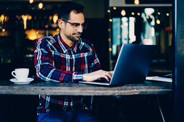 Concentrated young man copywriter in stylish eyeglasses keyboarding information on modern laptop computer sitting in coffee shop interior.Male graphic designer working freelance at digital netbook