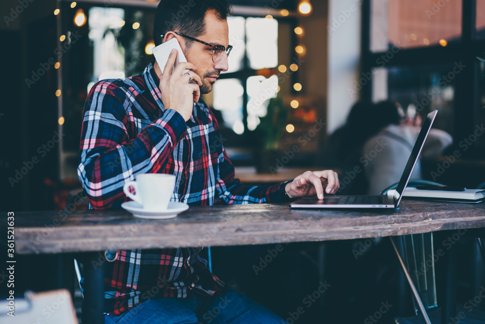 Poster side view of bearded man in eyeglasses making payment online on laptop and talking on smartphone to 