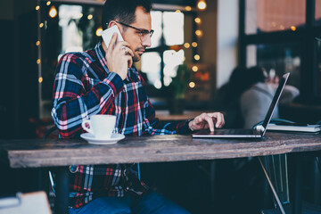 Side view of bearded man in eyeglasses making payment online on laptop and talking on smartphone to booking tickets.Hipster guy transferring money using internet on computer and calling to friend