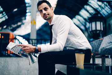 Young handsome professional employer dressed in formalwear looking away while sitting at bench outdoors.Positive entrepreneur in white shirt spending time outside with newspaper about finance