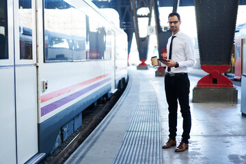 Handsome formally dressed male entrepreneur in eyewear waiting for train on platform enjoying coffee to go, prosperous businessman browsing information about public transport schedule getting to work