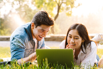 Image of couple using laptop and smiling while lying on grass