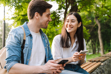 Image of pleased couple using mobile phones while sitting on bench