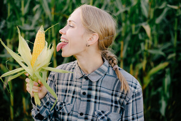 Beuatiful positive blonde girl in corn field with maze in hands. Girl licking corn by tip of tongue Beautiful girl tasting checking corn maize. Girl with long longue