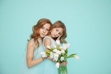 Two beautiful girls with curly locks of hair in dresses are embracing on blue background. Photo. White Tulip. Hair salon. Psychology of relationships. Look at camera. Friendship, love, family, touch