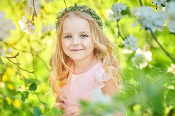 A beautiful little girl with a wreath on her head and a delicate pink dress plays in a blooming garden at sunset. Happy child resting outdoors