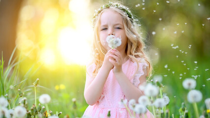 A beautiful little girl with a wreath on her head and a delicate pink dress plays with dandelions in the garden at sunset. Happy child resting outdoors