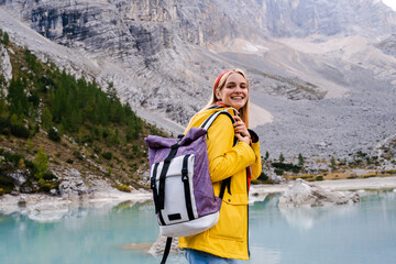 Lake Sorapis. Dolomites Alps. Italy. Blonde girl in yellow raincoat with backpack stays in the middle of azure water & smiles on background of pine forest & rock mountains