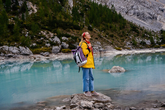 Lake Sorapis. Dolomites Alps. Italy. Blonde girl in yellow raincoat with backpack stays on stone in the middle of azure water & looks far away on background of pine forest & rock mountains