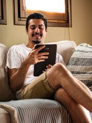 A picture of a brunette male sitting on a white sofa and holding a tablet