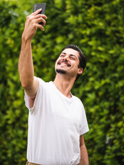 A vertical picture of a brunette male taking selfies against a greenery