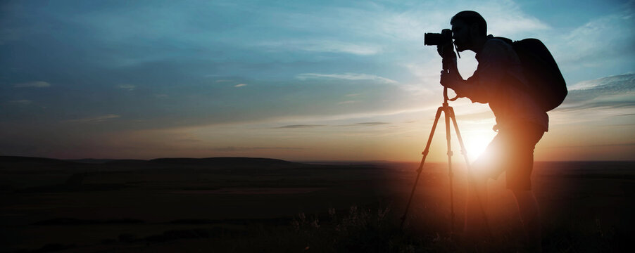 Side silhouette of photographer taking pictures using dslr camera and tripod on sunset or sunrise