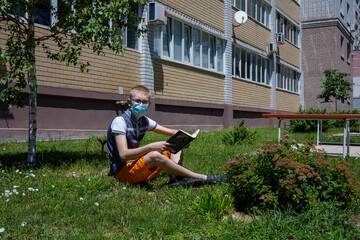 A boy in a protective mask and glasses reads a book on the green grass outside the house on a Sunny summer day. А new normal, new reality.
