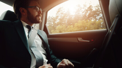 Low angle of businessman in classic suit traveling in car on back seat looking outside the window