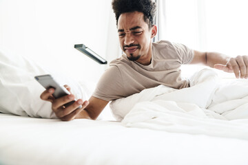 Fototapeta premium Photo of displeased african american man using mobile phone in white bed