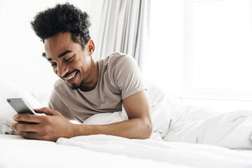 Photo of smiling african american man using mobile phone in white bed