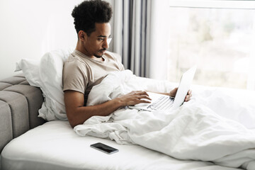 Photo of african american man working with laptop while lying in bed