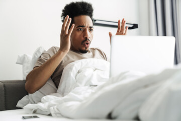Photo of african american man working with laptop while lying in bed