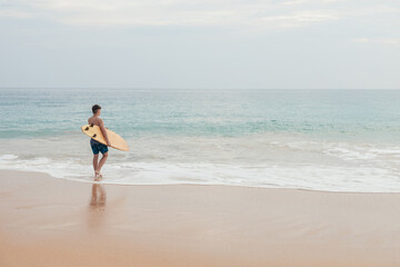 Handsome teenage boy with surfboard going into the ocean at sandy beach on surf line.
