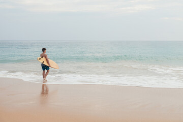 Fototapeta na wymiar Handsome teenage boy with surfboard going into the ocean at sandy beach on surf line.