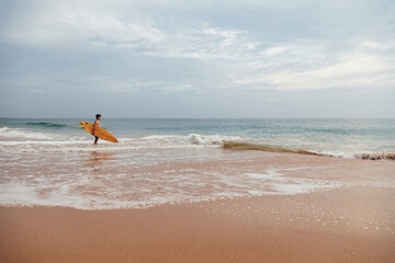 Fototapeta na wymiar Handsome teenage boy with surfboard going into the ocean at sandy beach on foamy surf line.