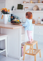 curious infant baby girl trying to reach things on the table in the kitchen with the help of step stool