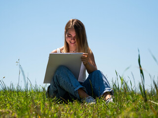 A beautiful female sketching on her notebook while sitting on a grass-covered field on a sunny day