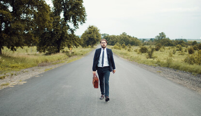 Businessman in classic suit holding briefcase moving forward in middle of country road looking...