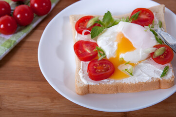 A delicious breakfast with poached egg, bread, cherry tomatoes, cheese and herbs. Poached egg for breakfast. Sandwich with cheese, red cherry tomatoes, herbs and poached egg.