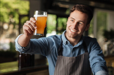 Quality fresh drink. Satisfied smiling barman in apron raises glass of light ale up and looks on its color