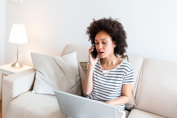 woman sitting on sofa with laptop and talking on phone at home. Young successful businesswoman working from home while talking at phone. College student studying on laptop and using phone.