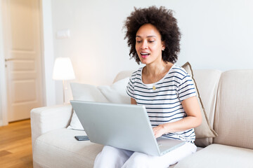 Young beautiful woman with curly hair working on laptop computer while sitting on the sofa at home, heck on oline shops for cyber monday sales. Technology woman concept alternative office freelance