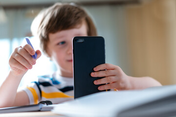 Back to school. Distance learning online education. Caucasian smile kid boy studying at home with smartphone and doing school homework. Thinking child siting at table with cell phone