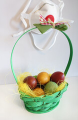 Green basket with colorful painted eggs and anti virus mask in background, hanging on white wall. Stay at home. Corona during easter time.