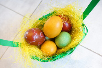 Colorful painted eggs in yellow nest in green basket top view. Happy Easter, stay at home!